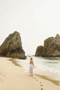 A person walking on a sandy beach towards two large rocks, with clear footprints behind them. The sky is slightly overcast.